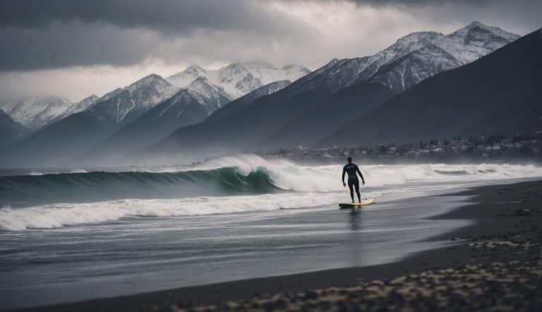 A lone surfer braves the winter waves, with snow-capped mountains in the background and a cold, grey sky overhead